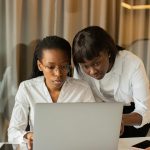 Two women in business attire working together on a laptop, fostering teamwork and cooperation.